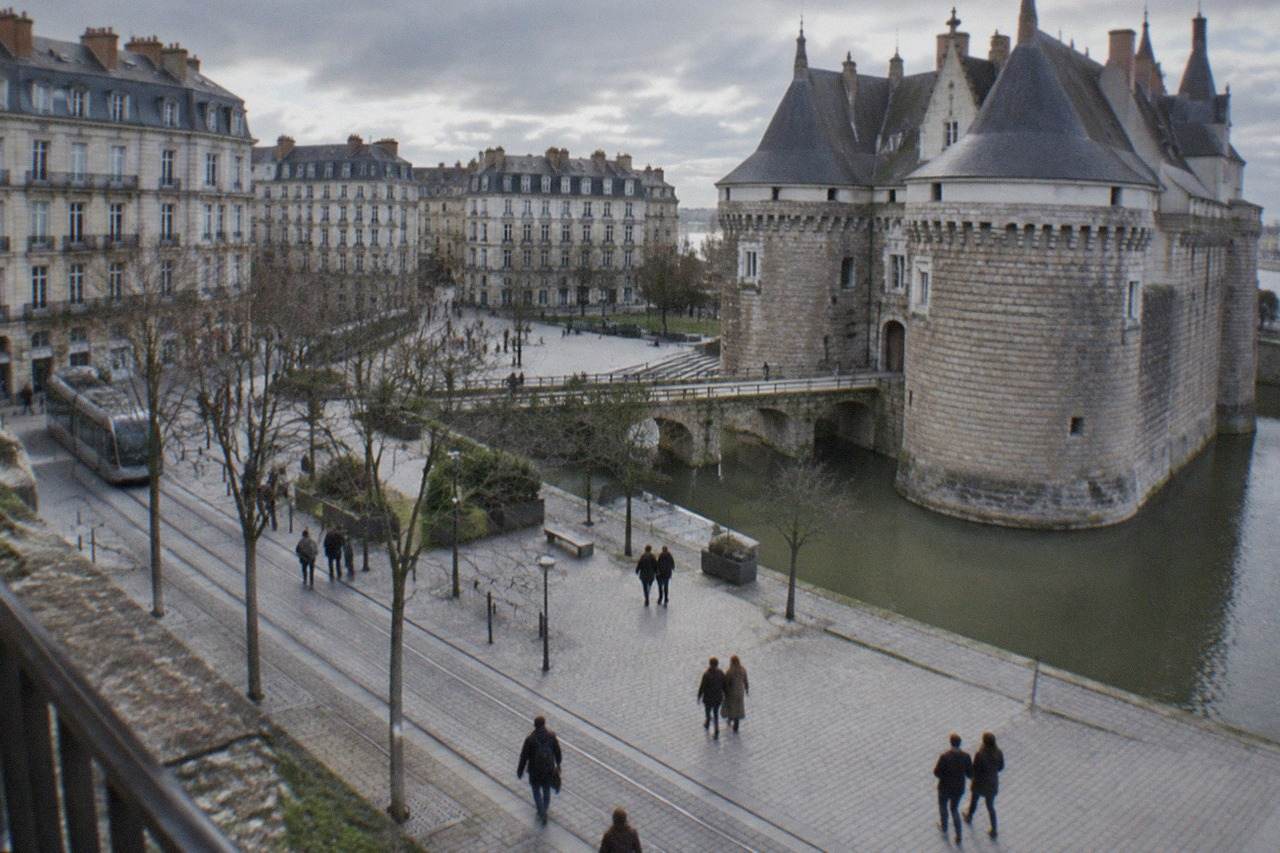 A stone castle with towers and a moat beside a plaza in a French city, people walking past on a mild day