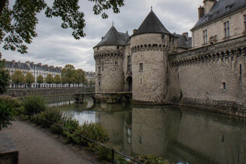 A medieval stone gateway and towers of a castle reflected in a water moat, seen from a garden path