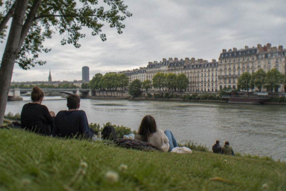 A group of travellers relaxing on a grassy riverbank beside the water in Nantes with trees and city buildings in the background