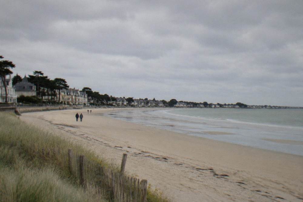 A sandy Atlantic beach at low tide with dune grass and distant seaside rooftops under a wide sky