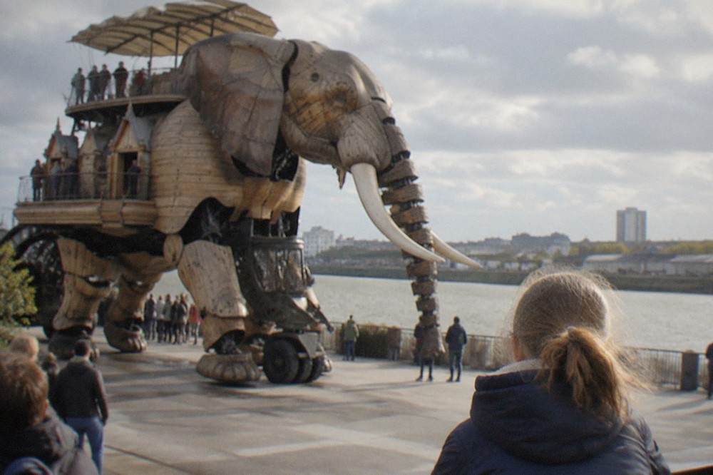 A child watching a large mechanical elephant sculpture move along a waterfront promenade