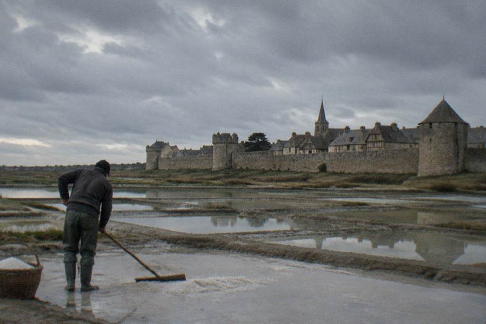 A salt worker raking grey crystals from a shallow clay pan with a long wooden tool, fleur de sel in a basket nearby