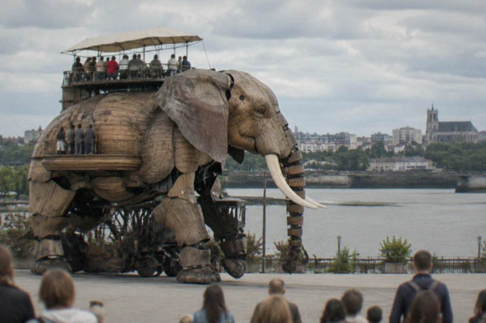 Passengers riding on the open terrace on the back of the wooden mechanical elephant as it walks beside the Loire