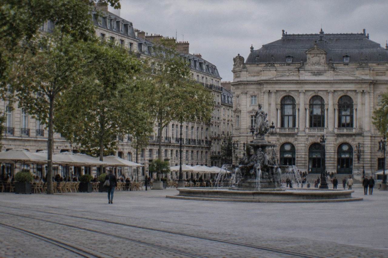 A grand stone square in Nantes with an ornate theatre facade, a central fountain and café terraces under plane trees