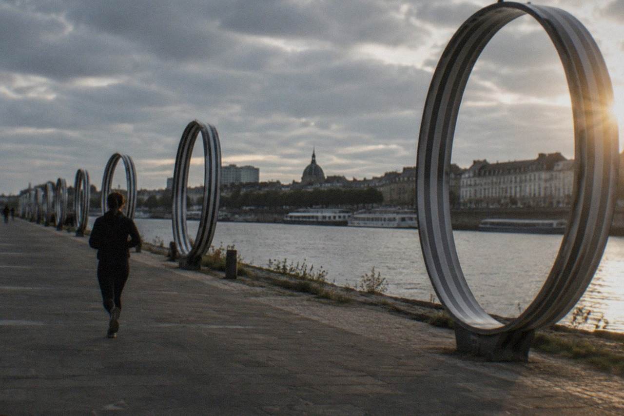 A row of striped rings standing along a riverside promenade with boats and a city skyline behind them