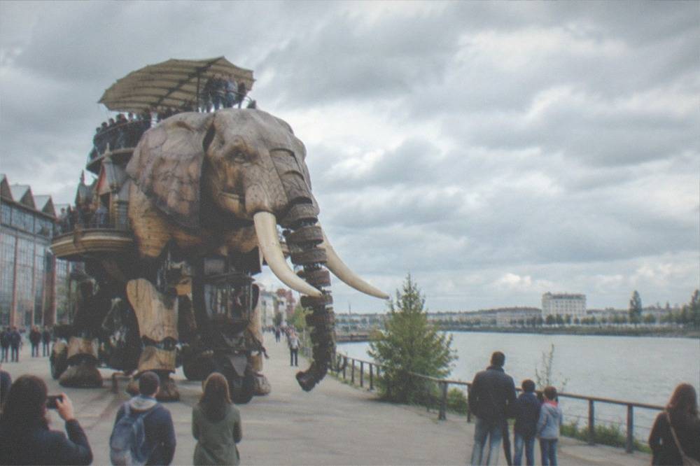 A mechanical elephant carrying visitors along a waterfront between warehouse buildings