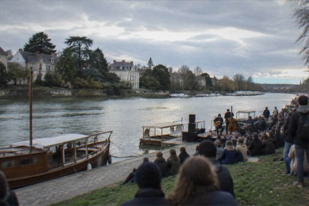 Crowds on a riverside quay listening to an open-air concert beside moored wooden boats