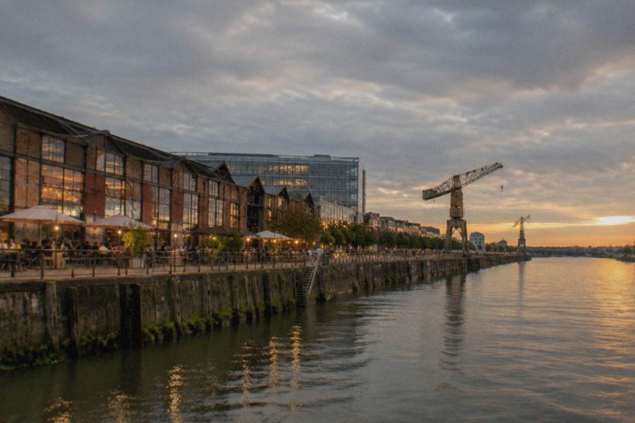 Long riverside warehouse on the west quay of the Île de Nantes at dusk, with lit bar terraces facing the Loire