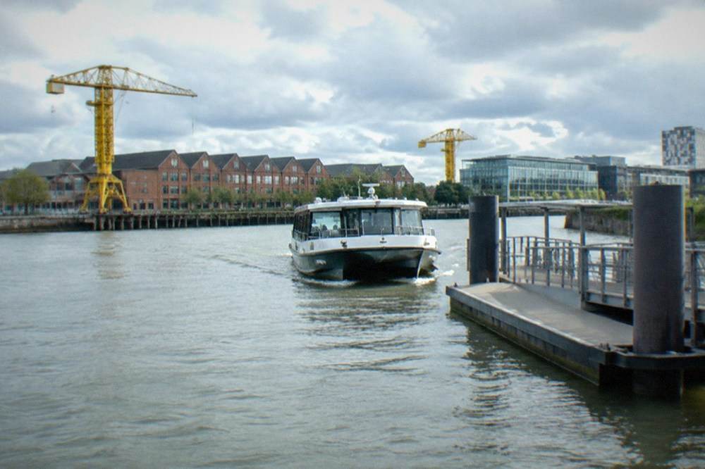 A river shuttle boat approaching a pontoon on the Loire, with the warehouse roofs of the Île de Nantes behind