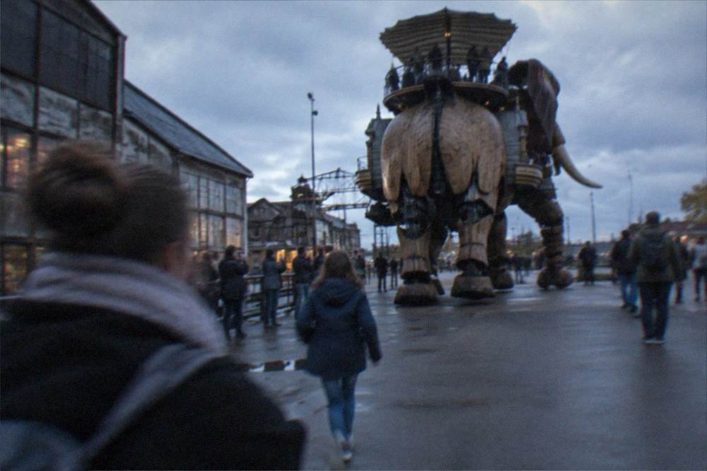 The mechanical elephant walking past converted shipyard buildings on the Île de Nantes, with visitors watching