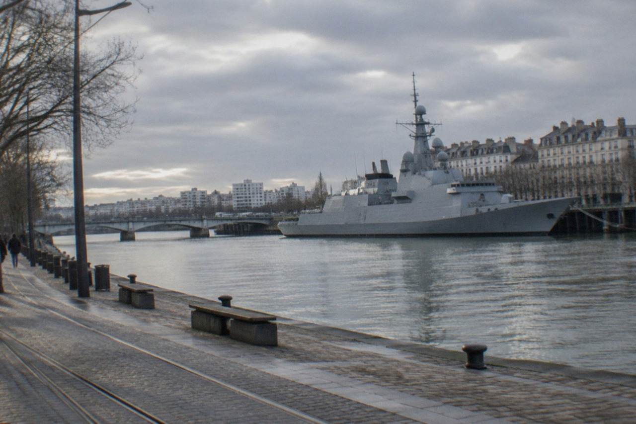 A grey naval destroyer moored on the Loire river at Nantes with apartment buildings and a bridge behind it