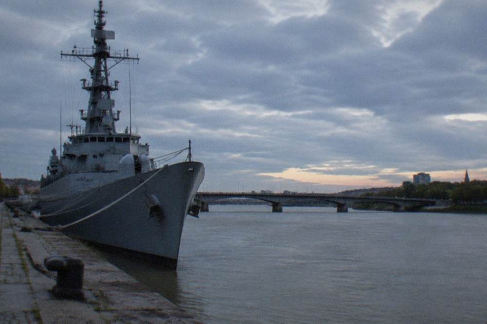 The grey bow of a destroyer seen from a stone quay with the Loire and a bridge behind it