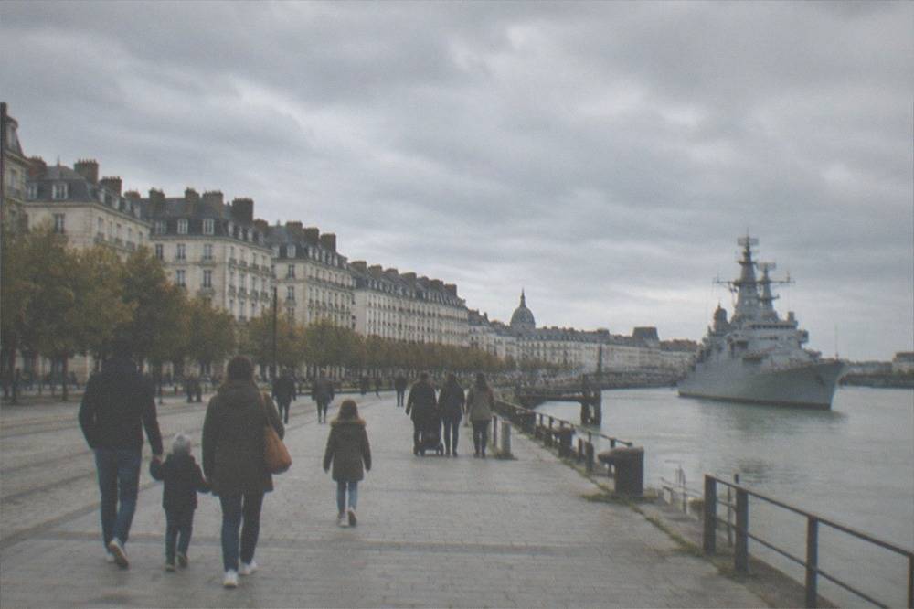 Families walking along a wide Loire-side quay in Nantes with a grey warship moored in the background