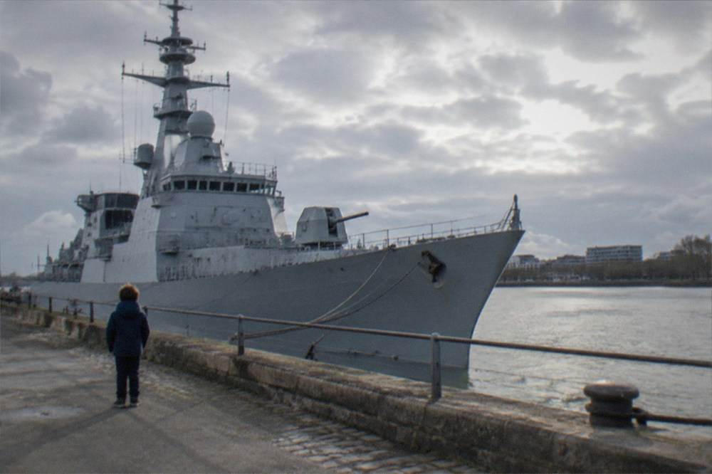 A child looking up at the tall grey superstructure of a moored destroyer from a riverside quay