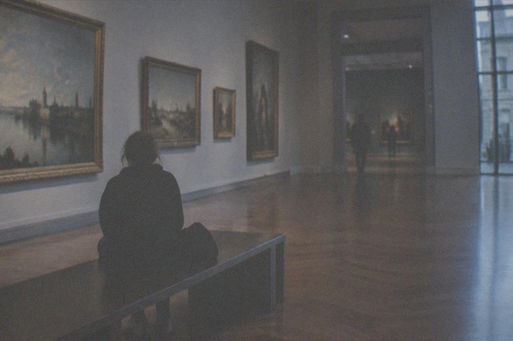Visitor resting on a bench in a long museum gallery lined with paintings