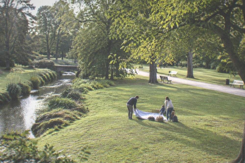 A family spreading a picnic blanket on a park lawn under trees
