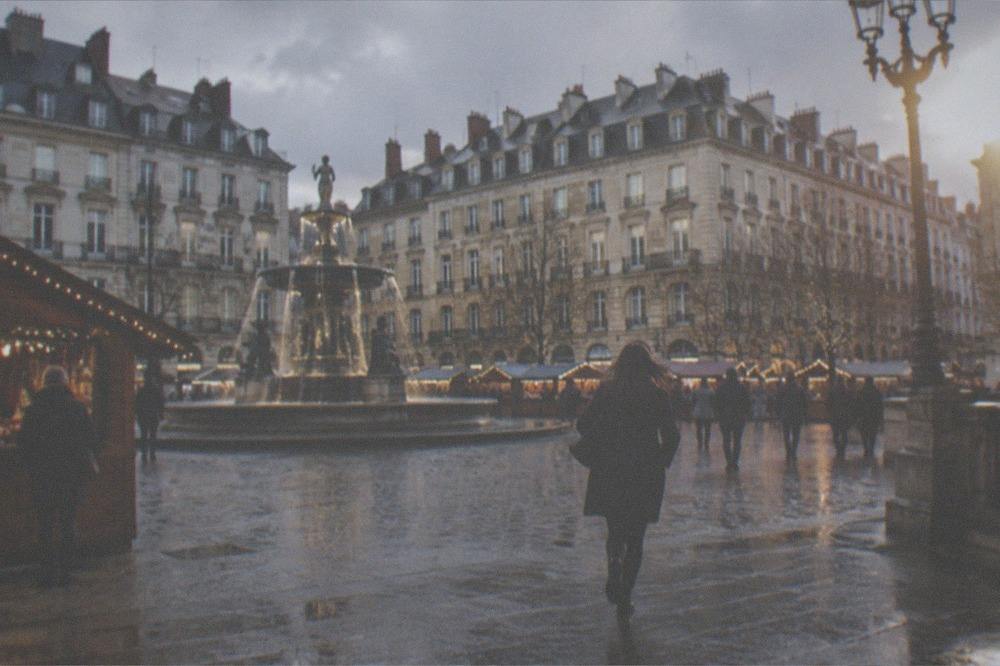 A European city square in December with wooden market chalets, strings of lights and an illuminated fountain
