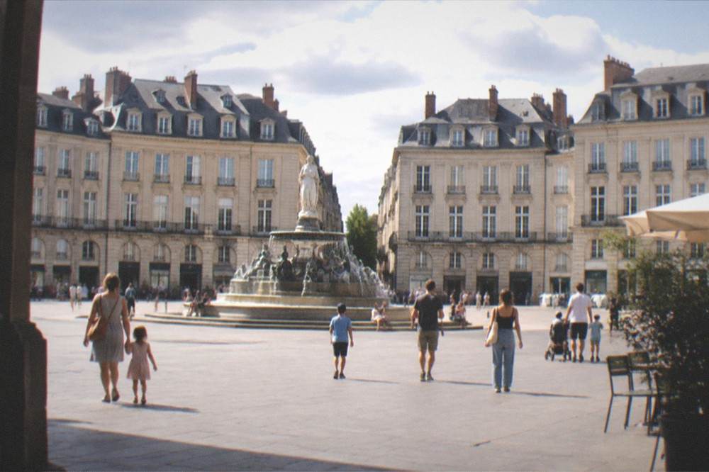 Families and children walking through a European city square with a fountain and classical buildings on a summer afternoon