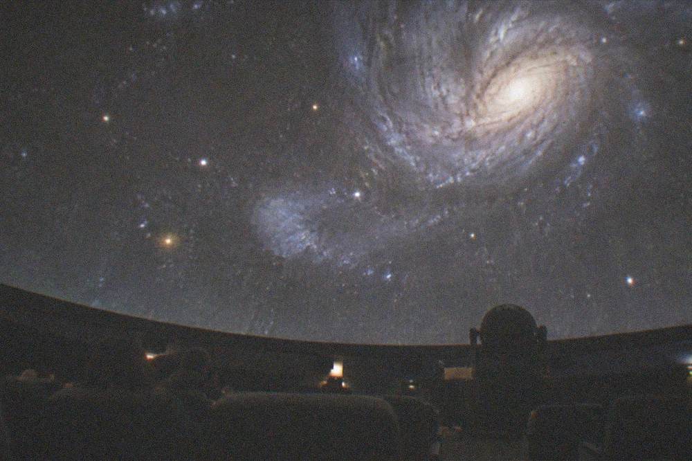A projected starfield and swirling galaxy filling the curved interior of a planetarium dome in near darkness