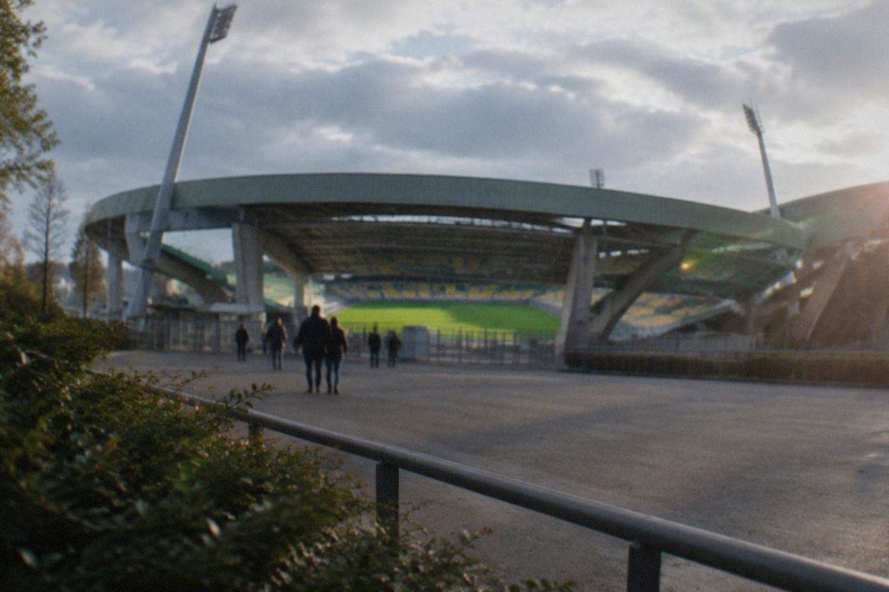 A large football stadium with curved stands and a bright green pitch seen from outside on a sunny day