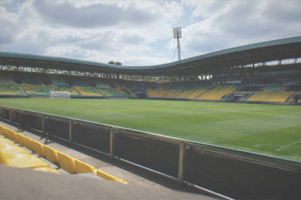A green football pitch and empty stands viewed from inside a stadium on a bright afternoon