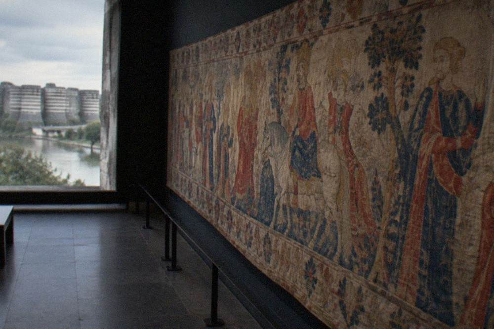 Detail of a long medieval wall-hanging hung in a dim gallery, woven figures in faded blues and reds