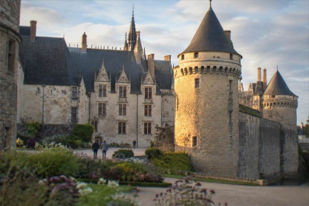 A walled castle courtyard with flower beds and stone towers under a blue sky with light clouds
