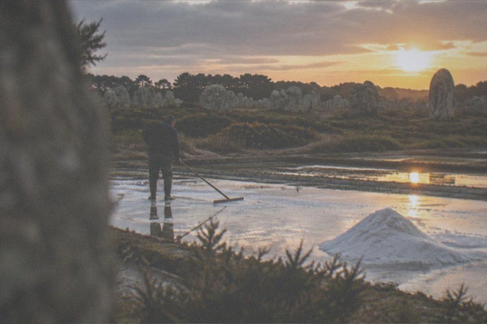 Salt marsh worker raking white salt crystals from a shallow rectangular pond at sunset