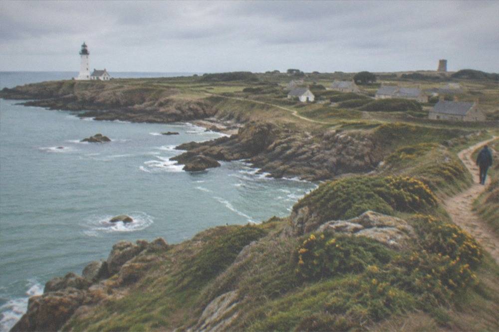 A path running along a low clifftop above turquoise sea, with a stone tower visible in the distance