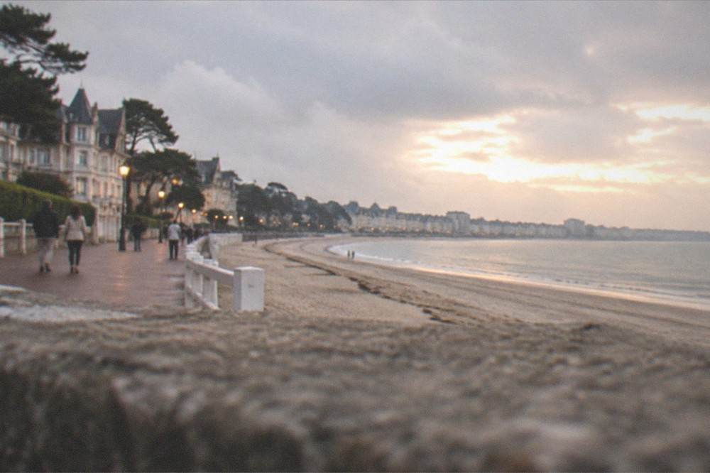 A seafront promenade at dusk with warm lighting, a few strollers and the bay beyond
