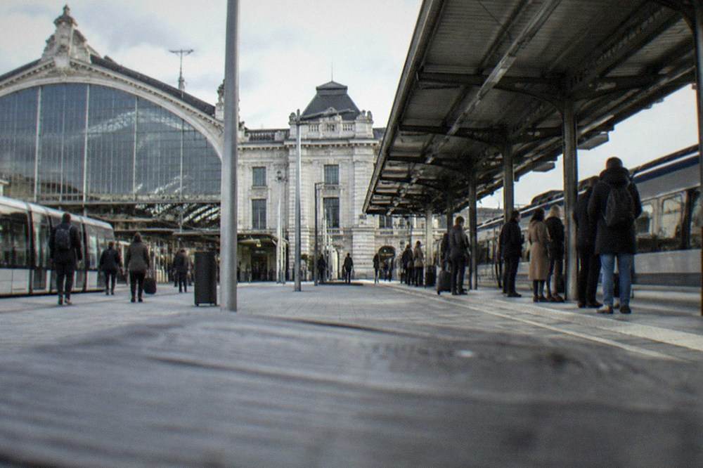 A regional train platform beside a French station building with passengers waiting with bags