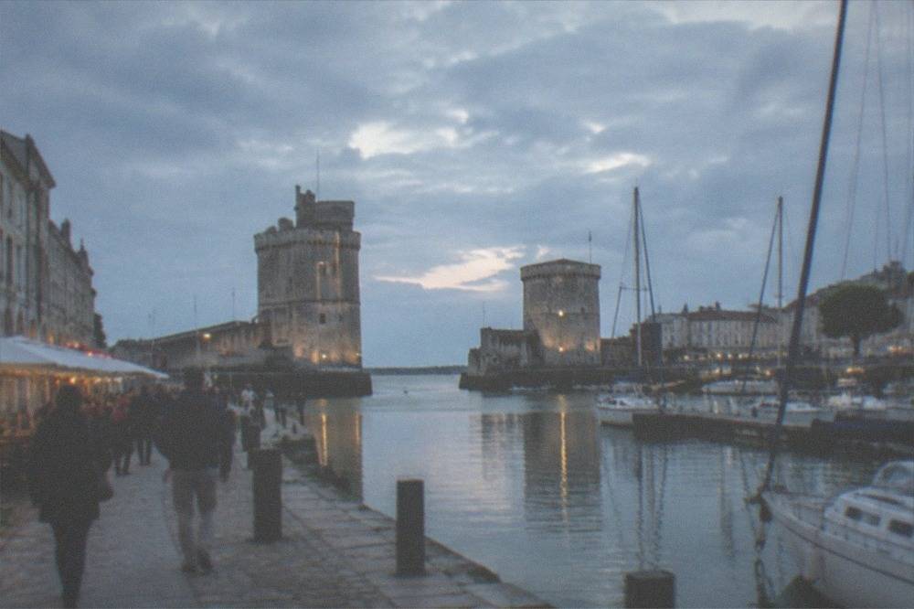 People strolling along a stone quay past cafes with moored sailing boats and historic towers behind