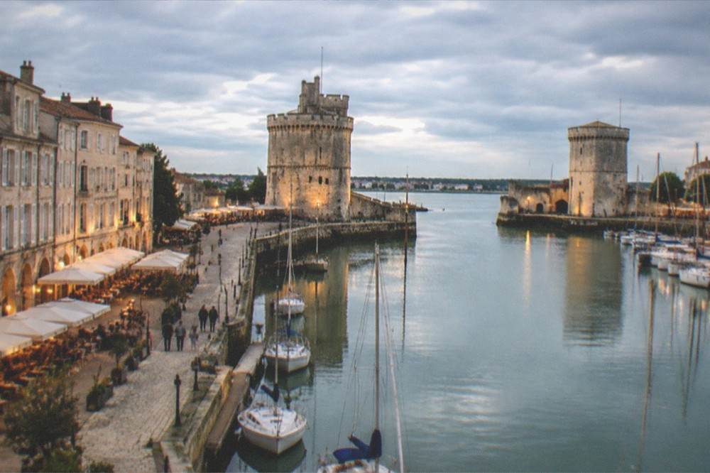 An evening harbour scene with lit cafes along a quay and boats reflected in still water