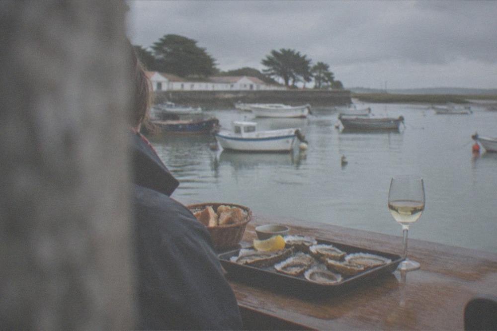 A tray of fresh open oysters on a wooden table beside a fishing harbour with small boats moored behind