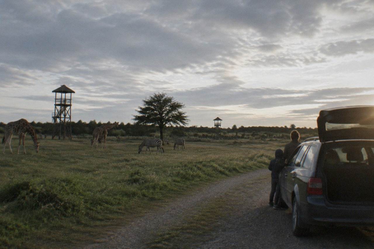 A family car parked on a gravel track beside open grassland where tall wooden observation posts stand against a wide sky