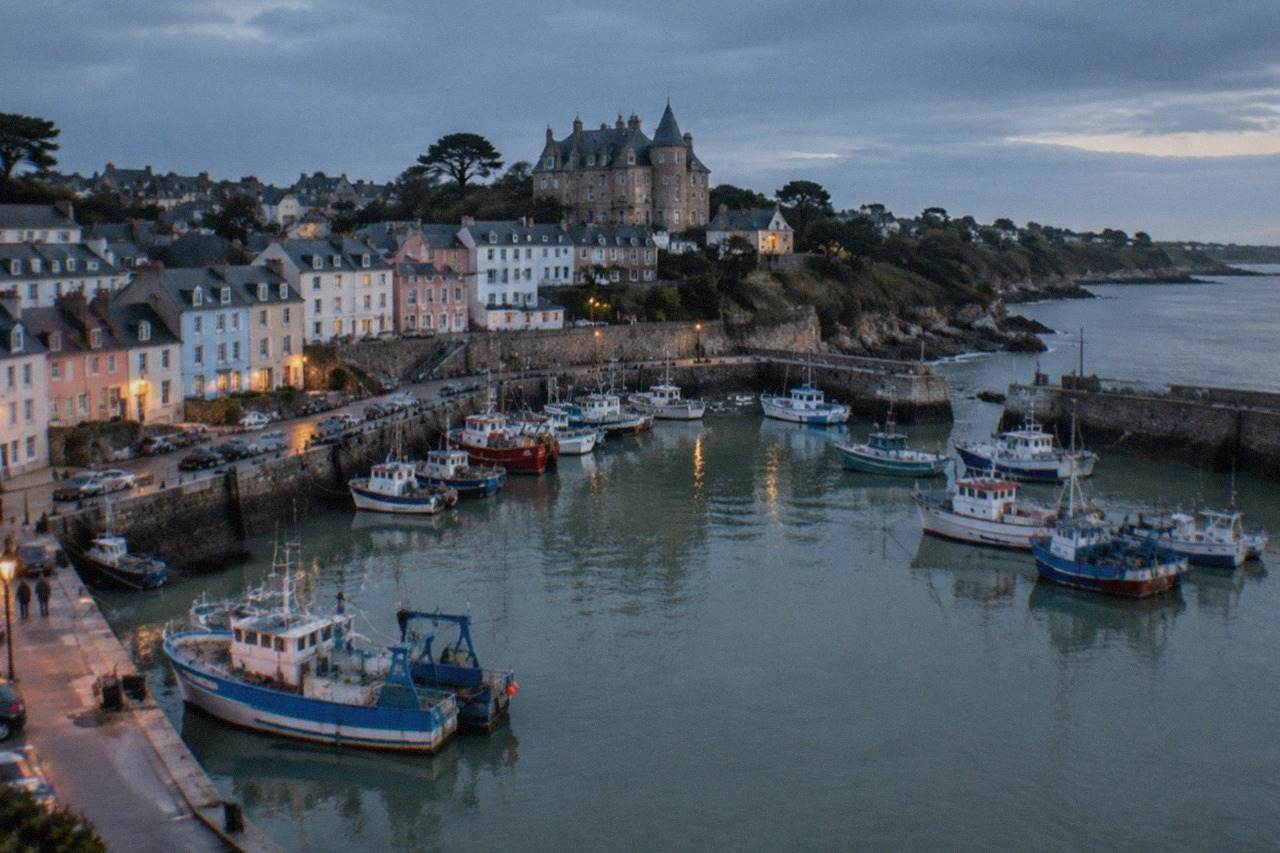 A small Atlantic seaside town seen from above, with a sheltered harbour of moored fishing boats and pastel houses rising behind it