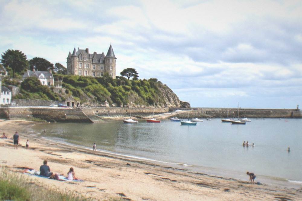 A calm bay beach at midday with families on the sand and a stone breakwater closing the far side of the cove