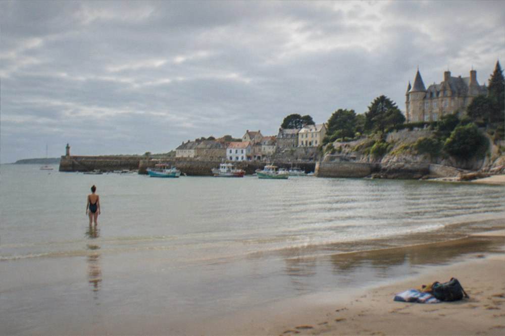 A swimmer standing in shallow seawater on a wide empty beach, with a towel and bag on the sand behind