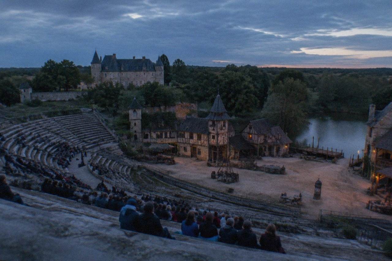 A large open-air amphitheatre with tiered stone seating facing a medieval village stage set
