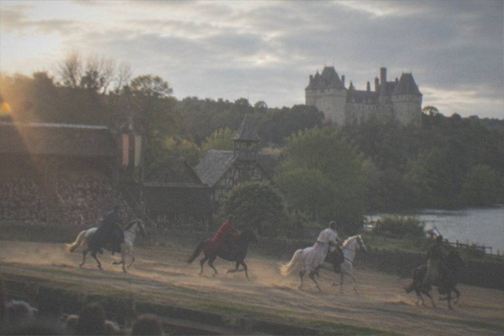 Costumed performers on horseback galloping across a dusty arena in front of a grandstand