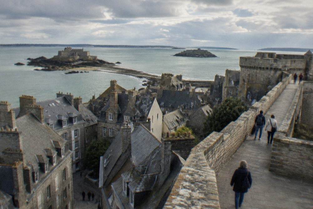 Walkers strolling along a stone rampart path with the sea and fortified islets beyond
