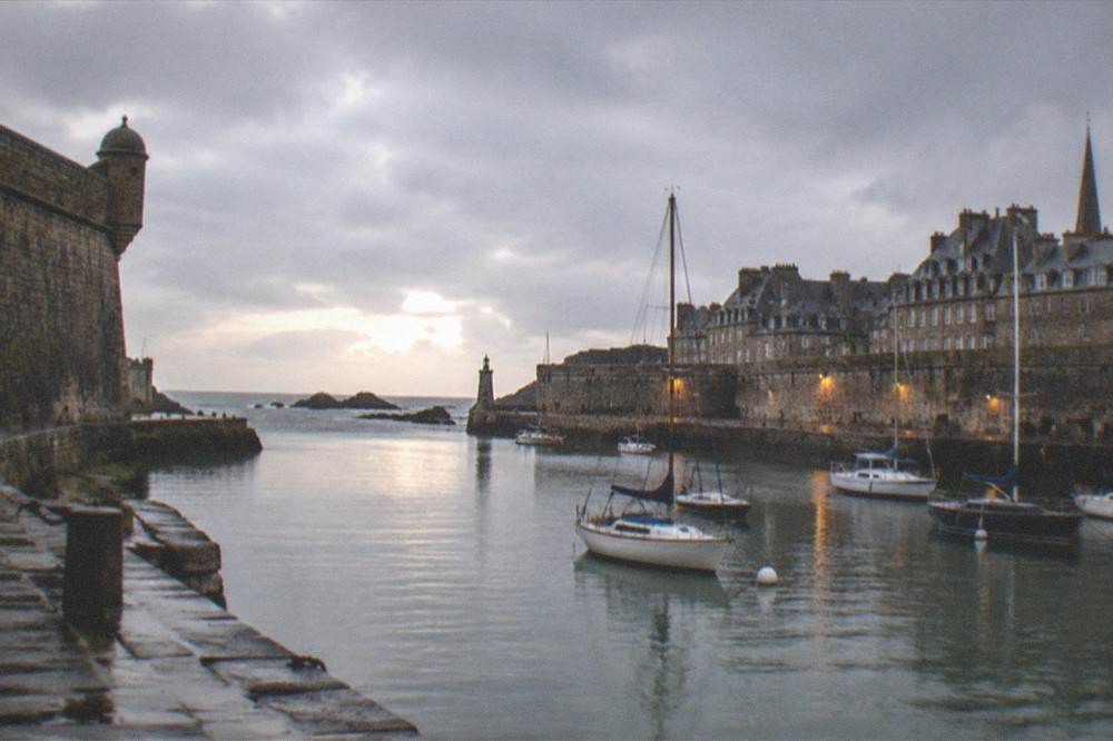 Harbour entrance of a walled port town with moored sailing boats and evening light on stone defences