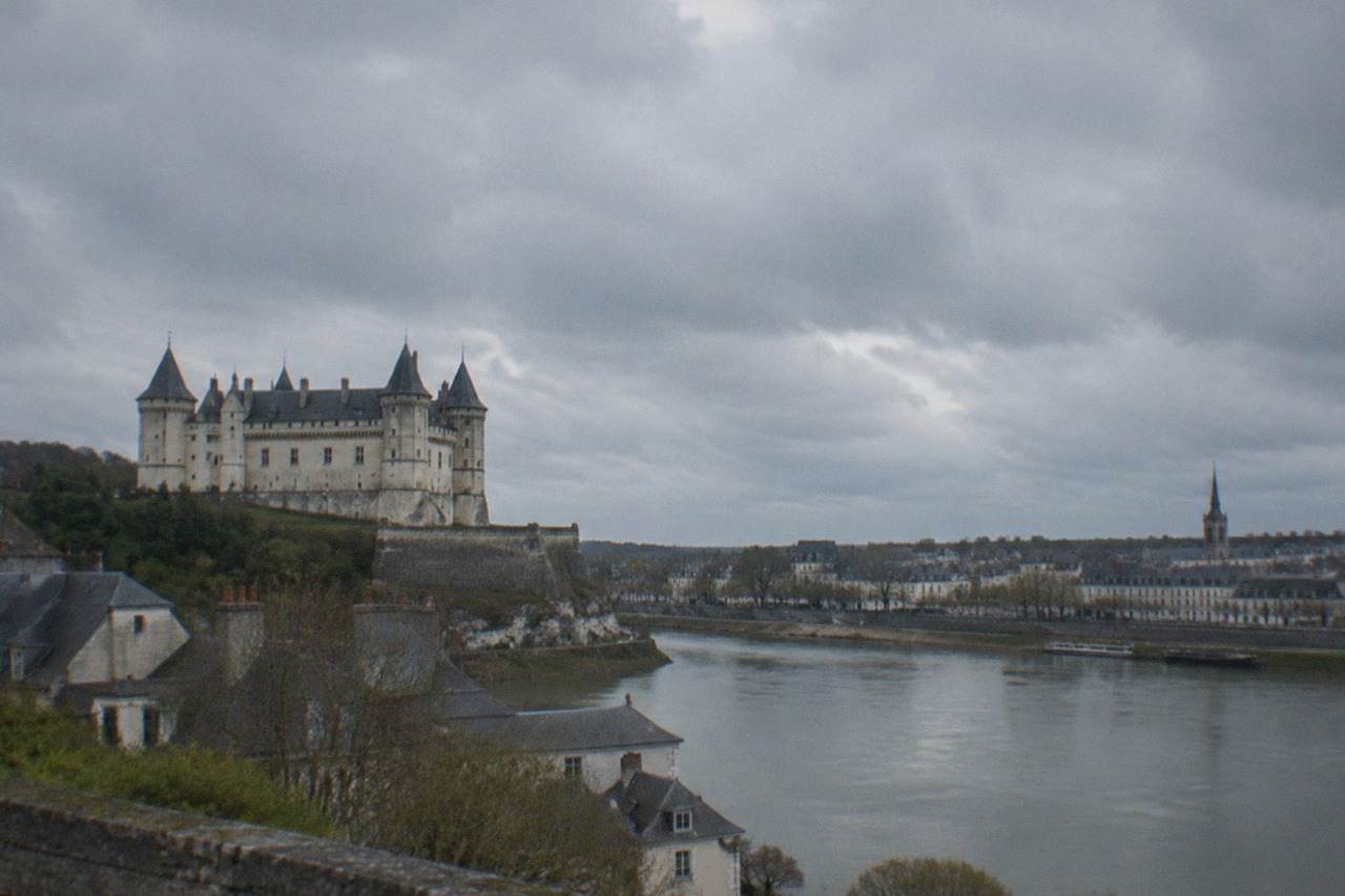 A pale stone château with round towers standing above a wide river, with a town of grey roofs on the far bank