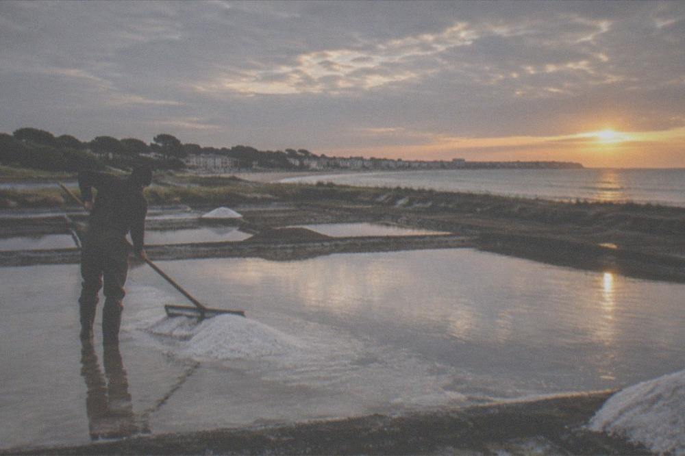Salt marsh worker raking white sea salt from shallow square pans at sunset