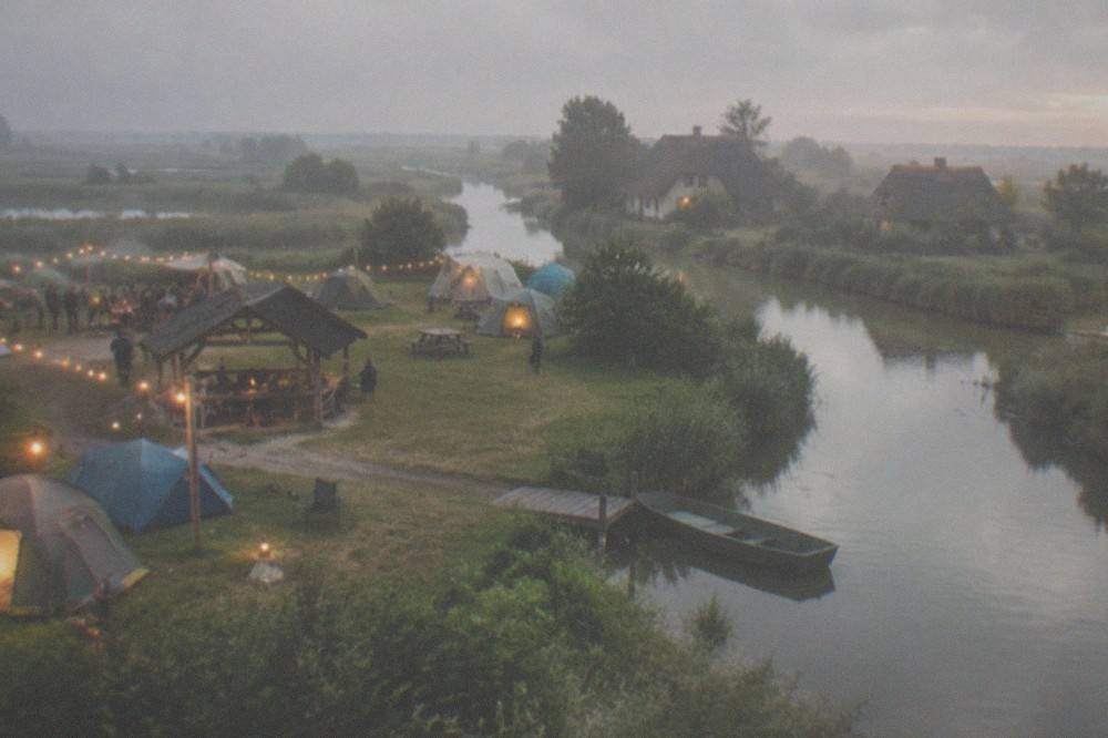 Dusk over a campsite field with strings of warm lights, a communal barbecue shelter and tents glowing in the evening