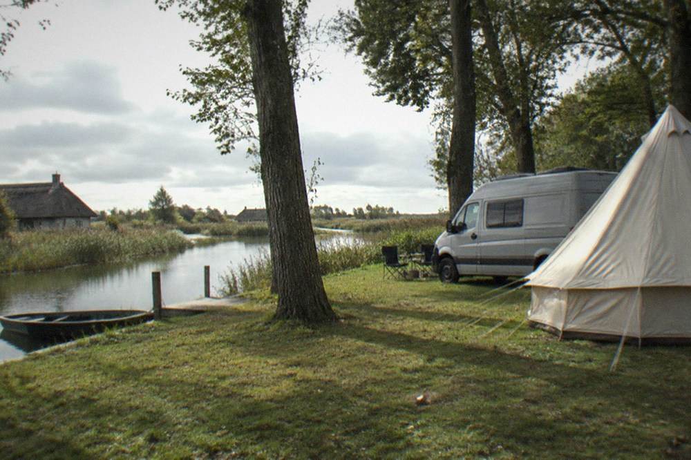 A campsite pitch under tall shade trees in late afternoon, a campervan and a canvas bell tent on mown grass