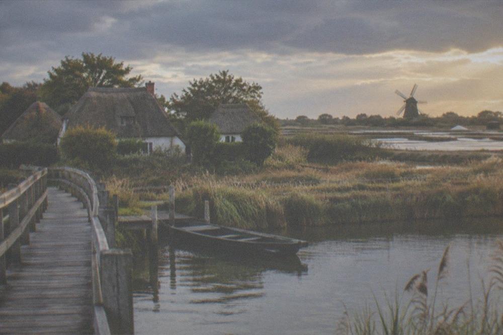 A wooden walkway over a salt marsh at golden hour, a traditional salt pan and windmill in the distance