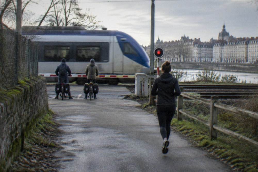Two cyclists with panniers waiting at a rural level crossing while a regional train passes on the line to Nantes