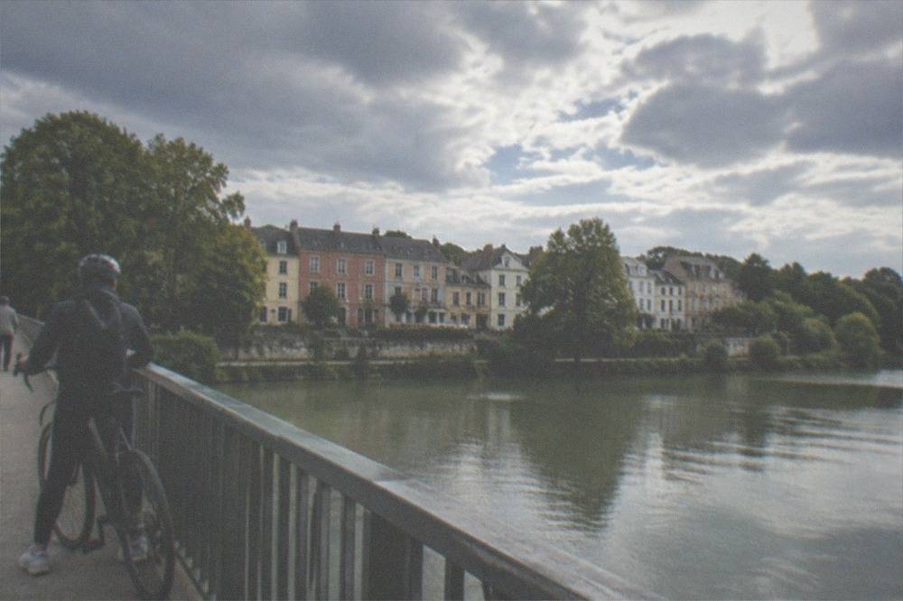 A cyclist pausing on a bridge over a river with colourful cottage facades on the far bank