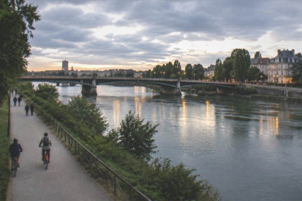 Two cyclists crossing a wide bridge at dusk with city lights reflecting on the river below